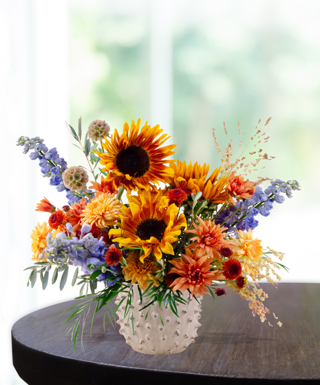 Bright fall floral arrangement with yellow sunflowers, orange mums, blue delphinium, and rustic accents in a white hobnail vase