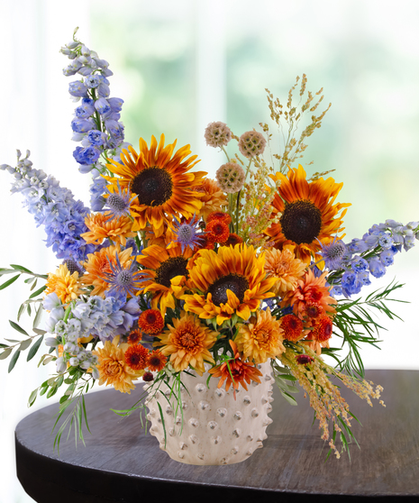 Bright fall floral arrangement with yellow sunflowers, orange mums, blue delphinium, and rustic accents in a white hobnail vase
