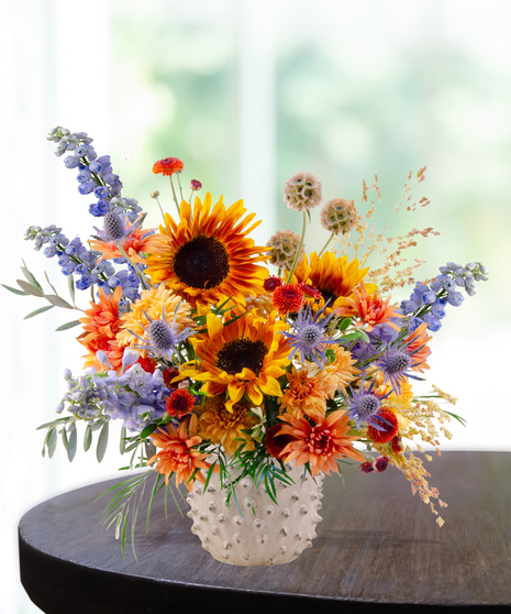 Bright fall floral arrangement with yellow sunflowers, orange mums, blue delphinium, and rustic accents in a white hobnail vase