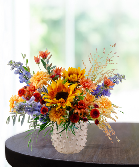 Bright fall floral arrangement with yellow sunflowers, orange mums, blue delphinium, and rustic accents in a white hobnail vase