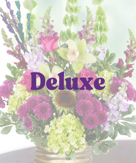 A Mardi Gras-themed arrangement featuring hot pink roses, green cymbidium orchids, purple mums, hydrangeas, and bells of Ireland in a gold vase with colorful bead accents.