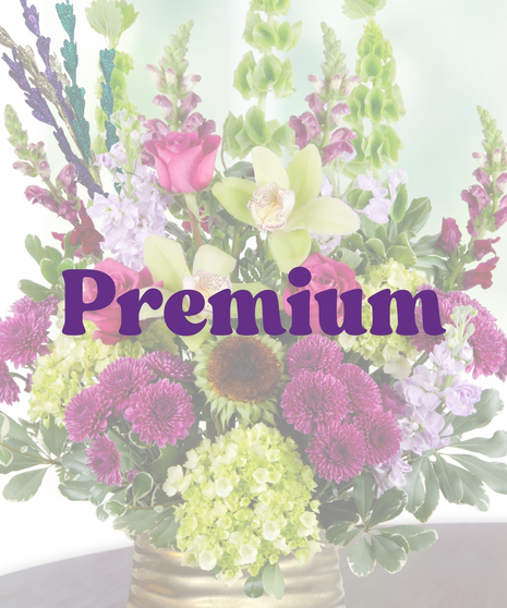A Mardi Gras-themed arrangement featuring hot pink roses, green cymbidium orchids, purple mums, hydrangeas, and bells of Ireland in a gold vase with colorful bead accents.