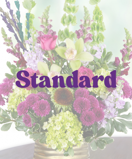 A Mardi Gras-themed arrangement featuring hot pink roses, green cymbidium orchids, purple mums, hydrangeas, and bells of Ireland in a gold vase with colorful bead accents.