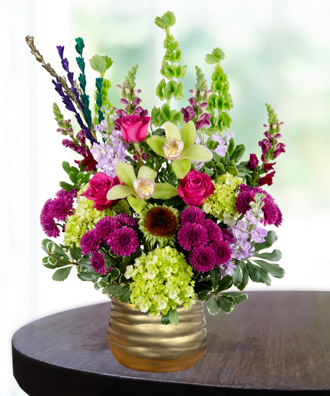 A Mardi Gras-themed arrangement featuring hot pink roses, green cymbidium orchids, purple mums, hydrangeas, and bells of Ireland in a gold vase with colorful bead accents.