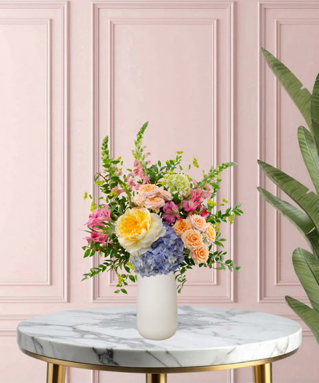 Because She's Mom Mother's Day flower arrangement featuring yellow garden roses, blue hydrangeas, pink snapdragons, and peach spray roses in a matte white vase.