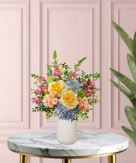 Because She's Mom Mother's Day flower arrangement featuring yellow garden roses, blue hydrangeas, pink snapdragons, and peach spray roses in a matte white vase.