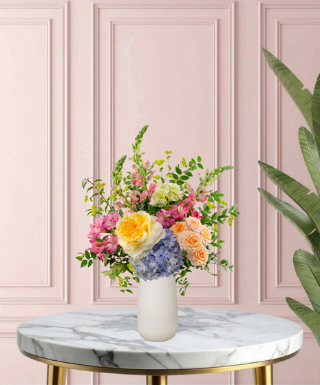 Because She's Mom Mother's Day flower arrangement featuring yellow garden roses, blue hydrangeas, pink snapdragons, and peach spray roses in a matte white vase.