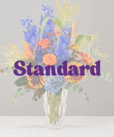 Bess fall flower arrangement with blue delphinium, sunflowers, Birds of Paradise, hydrangea, and orange roses in a crystal vase