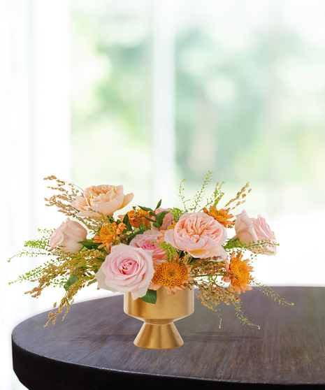 Boho-inspired floral arrangement with blush roses, peach garden roses, yellow mums, and greenery in a gold pedestal vase