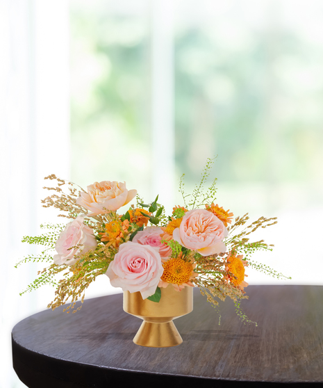 Boho-inspired floral arrangement with blush roses, peach garden roses, yellow mums, and greenery in a gold pedestal vase