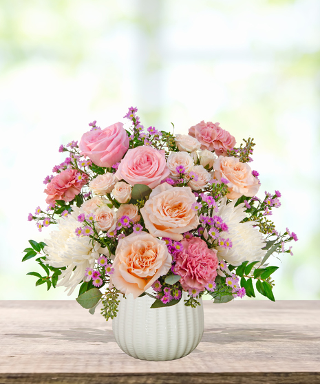 Peepin’ Pretty floral arrangement with blush and fuchsia roses, carnations, ivory mums, and lavender asters in a white ceramic vase.