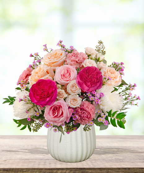 Peepin’ Pretty floral arrangement with blush and fuchsia roses, carnations, ivory mums, and lavender asters in a white ceramic vase.