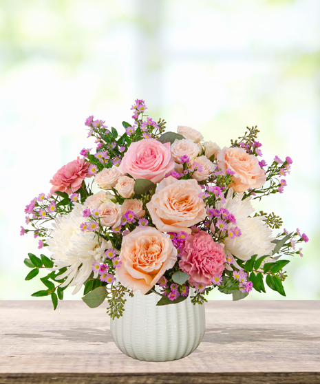 Peepin’ Pretty floral arrangement with blush and fuchsia roses, carnations, ivory mums, and lavender asters in a white ceramic vase.