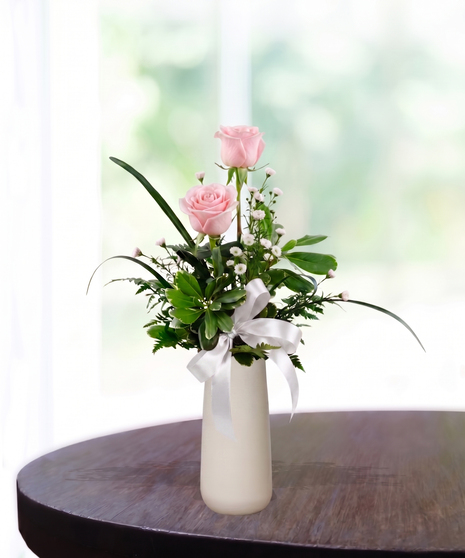 Bud Vase with six pink roses, white baby's breath, and a pink satin ribbon in a tall white ceramic vase.