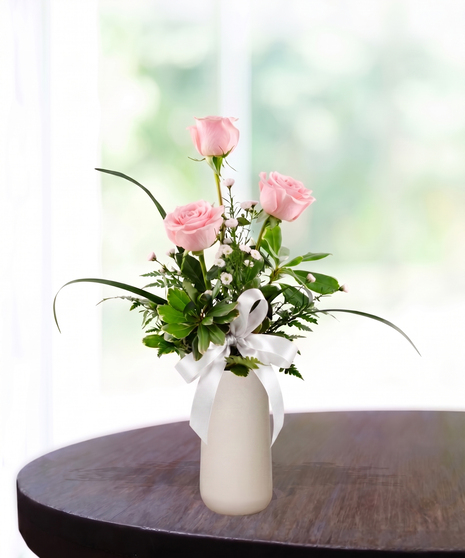 Bud Vase with six pink roses, white baby's breath, and a pink satin ribbon in a tall white ceramic vase.