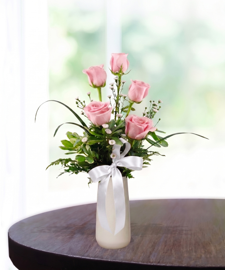 Bud Vase with six pink roses, white baby's breath, and a pink satin ribbon in a tall white ceramic vase.