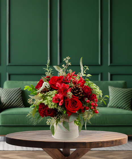 Cozy Cabernet rich red Christmas flower arrangement with deep red roses, green foliage, pine cones, and red berries in a textured vase