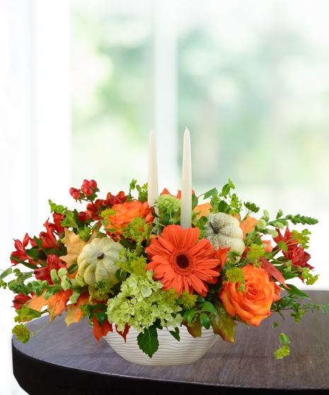 Fall floral centerpiece with orange gerbera daisies, roses, green hydrangeas, mini pumpkins, and candles in a ceramic bowl