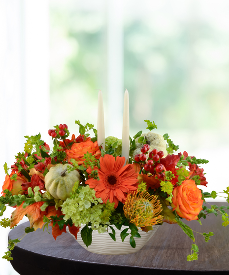 Fall floral centerpiece with orange gerbera daisies, roses, green hydrangeas, mini pumpkins, and candles in a ceramic bowl