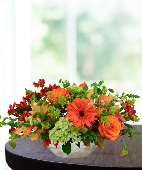 Fall floral centerpiece with orange gerbera daisies, roses, green hydrangeas, mini pumpkins, and candles in a ceramic bowl