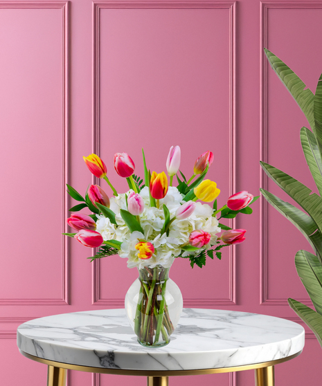 A vibrant bouquet of colorful tulips in yellow, orange, pink, and red, paired with white hydrangeas and greenery in a clear glass vase.