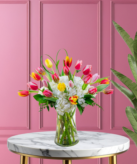 A vibrant bouquet of colorful tulips in yellow, orange, pink, and red, paired with white hydrangeas and greenery in a clear glass vase.