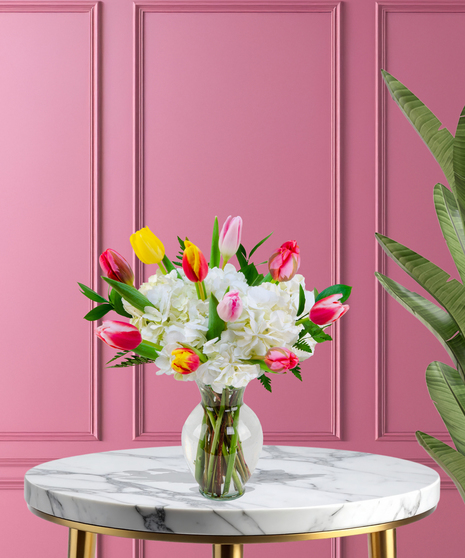 A vibrant bouquet of colorful tulips in yellow, orange, pink, and red, paired with white hydrangeas and greenery in a clear glass vase.