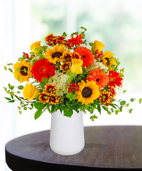 Bright fall flower arrangement with sunflowers, gerbera daisies, yellow roses, and bicolor mums in a white vase.