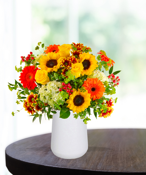 Bright fall flower arrangement with sunflowers, gerbera daisies, yellow roses, and bicolor mums in a white vase.