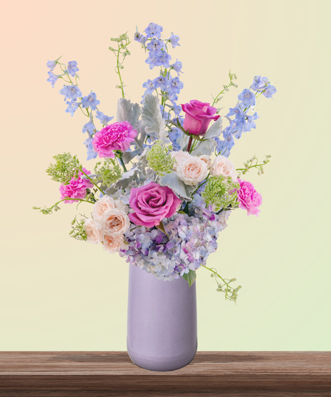 Mommalicious floral arrangement with pink roses, stargazer lilies, delphinium, lavender hydrangeas, and Queen Anne’s lace in a tall lavender vase – perfect for Mother’s Day.