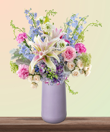 Mommalicious floral arrangement with pink roses, stargazer lilies, delphinium, lavender hydrangeas, and Queen Anne’s lace in a tall lavender vase – perfect for Mother’s Day.