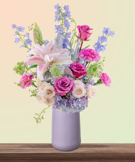 Mommalicious floral arrangement with pink roses, stargazer lilies, delphinium, lavender hydrangeas, and Queen Anne’s lace in a tall lavender vase – perfect for Mother’s Day.