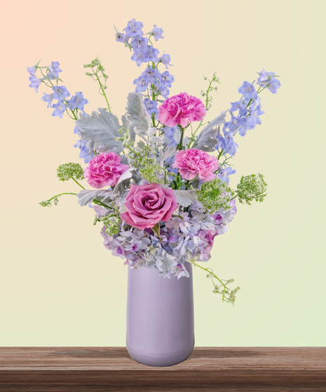Mommalicious floral arrangement with pink roses, stargazer lilies, delphinium, lavender hydrangeas, and Queen Anne’s lace in a tall lavender vase – perfect for Mother’s Day.