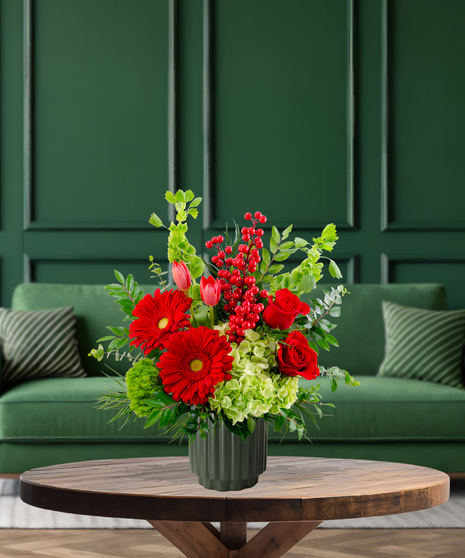 Deck the Halls vibrant Christmas arrangement with red gerbera daisies, red roses, pink tulips, green hydrangeas, Bells of Ireland, and red berries in a green textured vase