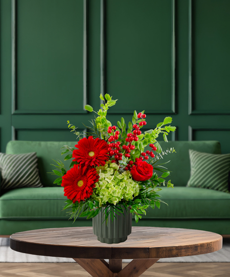 Deck the Halls vibrant Christmas arrangement with red gerbera daisies, red roses, pink tulips, green hydrangeas, Bells of Ireland, and red berries in a green textured vase
