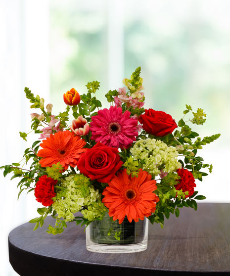Heatwave flower arrangement with red roses, orange gerberas, pink daisies, and pincushion protea in a clear glass cube vase