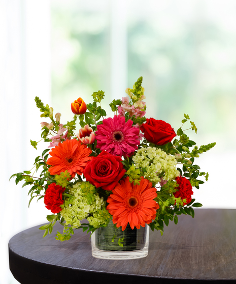 Heatwave flower arrangement with red roses, orange gerberas, pink daisies, and pincushion protea in a clear glass cube vase