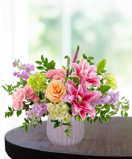 Spellbound flower arrangement with pink rose lilies, peach garden roses, pink spray roses, lavender stock, and green hydrangea in a soft blush vase