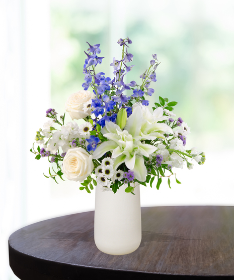 December Skies birthday flower arrangement with white roses, white lilies, blue delphinium, blue hydrangeas, white snapdragons, and green foliage in a frosted white vase