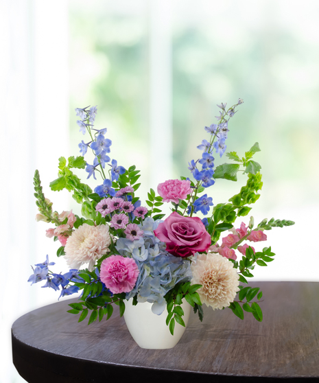Malibu Splash flower arrangement featuring magenta garden roses, lavender roses, blue hydrangeas, and blue delphinium in a matte white ceramic bowl.
