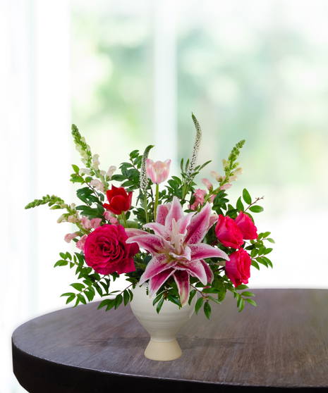 Legally Pink flower arrangement featuring hot pink roses, pink lilies, pink tulips, and white veronica in a matte cream ceramic pedestal vase.