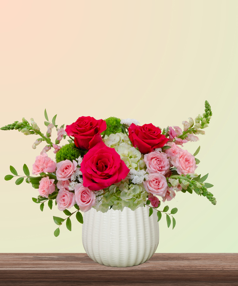 She’s All That! floral arrangement with hot pink roses, blush spray roses, green hydrangeas, snapdragons, and green dianthus in a white pedestal vase.