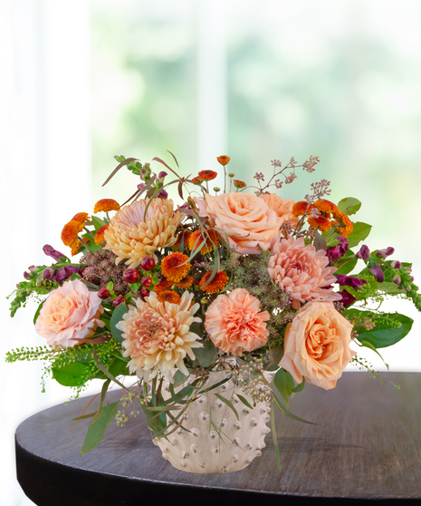 Peach, rust, and burgundy fall flower arrangement with roses, mums, snapdragons, and greenery in a white hobnail vase