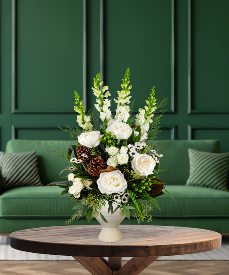 Evergreen Wonder flower arrangement with white roses, white spray roses, white snapdragons, evergreen foliage, green hypericum berries, and pine cones in a white vase