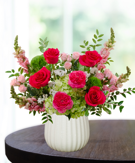 She’s All That! floral arrangement with hot pink roses, blush spray roses, green hydrangeas, snapdragons, and green dianthus in a white pedestal vase.