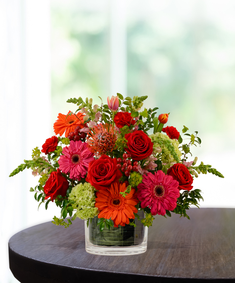 Heatwave flower arrangement with red roses, orange gerberas, pink daisies, and pincushion protea in a clear glass cube vase