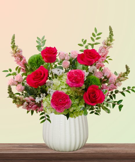 She’s All That! floral arrangement with hot pink roses, blush spray roses, green hydrangeas, snapdragons, and green dianthus in a white pedestal vase.