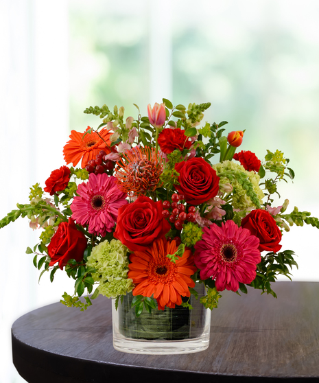 Heatwave flower arrangement with red roses, orange gerberas, pink daisies, and pincushion protea in a clear glass cube vase