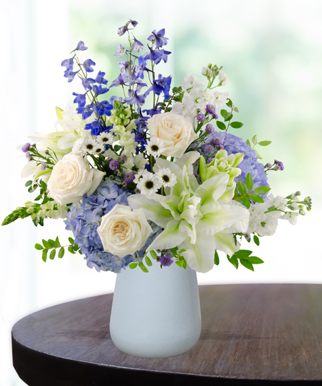 September birthday flower arrangement with blue hydrangeas, white roses, lilies, and delphinium in a soft blue vase.