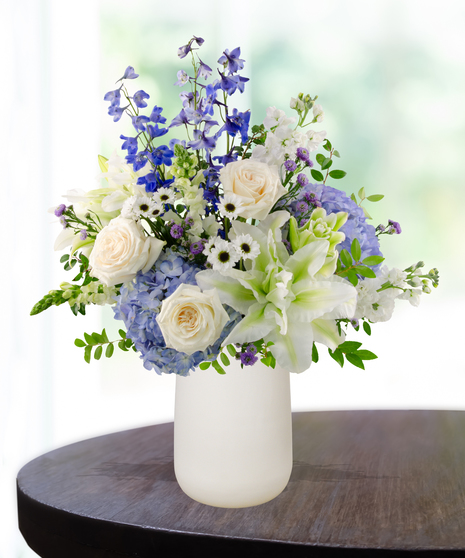 December Skies birthday flower arrangement with white roses, white lilies, blue delphinium, blue hydrangeas, white snapdragons, and green foliage in a frosted white vase